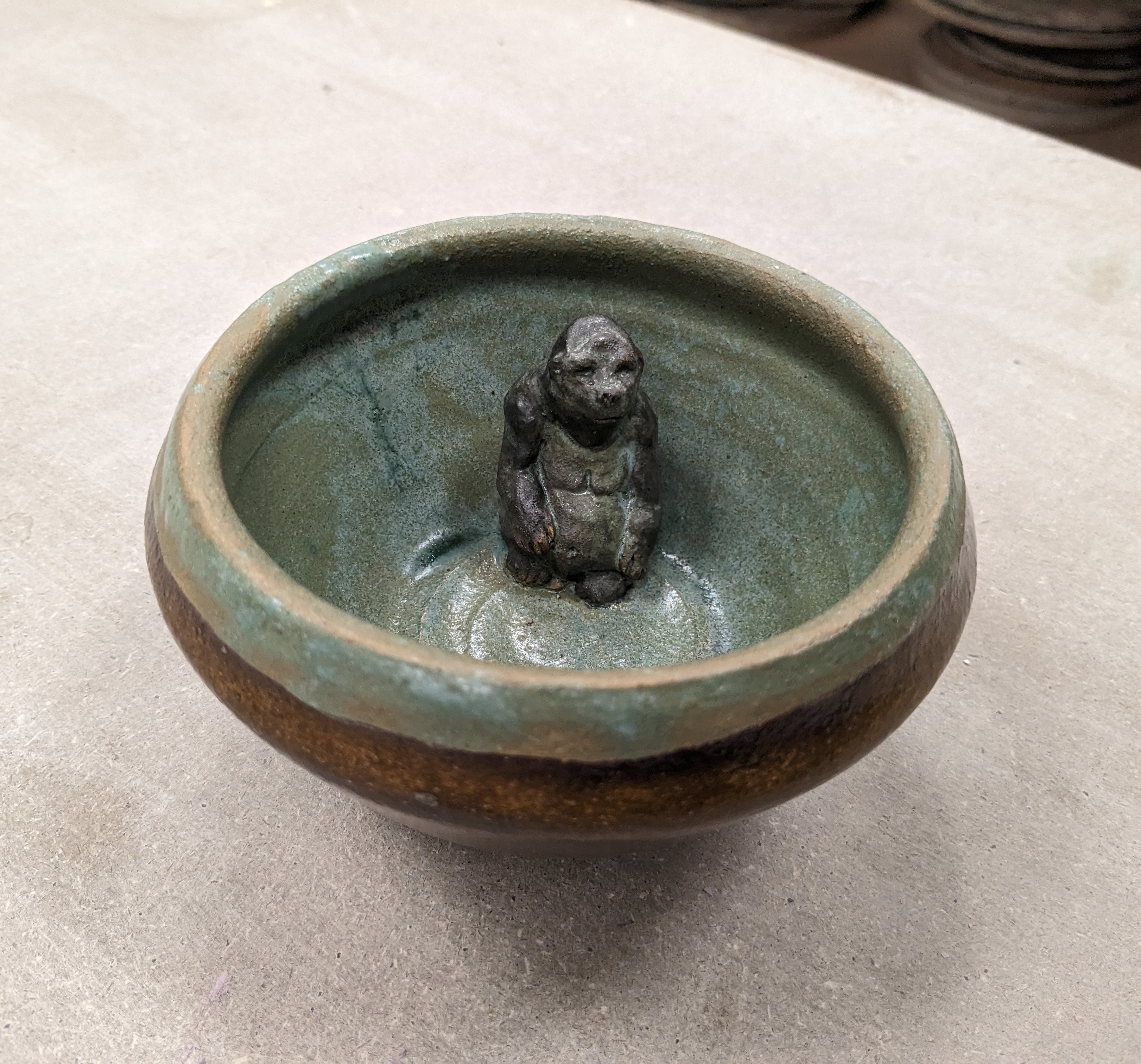 Ceramic bowl, brown on the outside and green on the inside, with a dark green ceramic gorilla sitting pensively inside the bowl